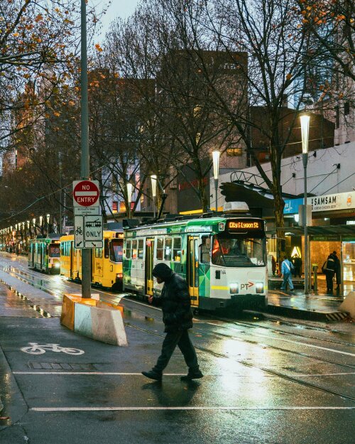 Pedestrian crosses a rainy street in Melbourne, with a bicycle lane and tram visible too.